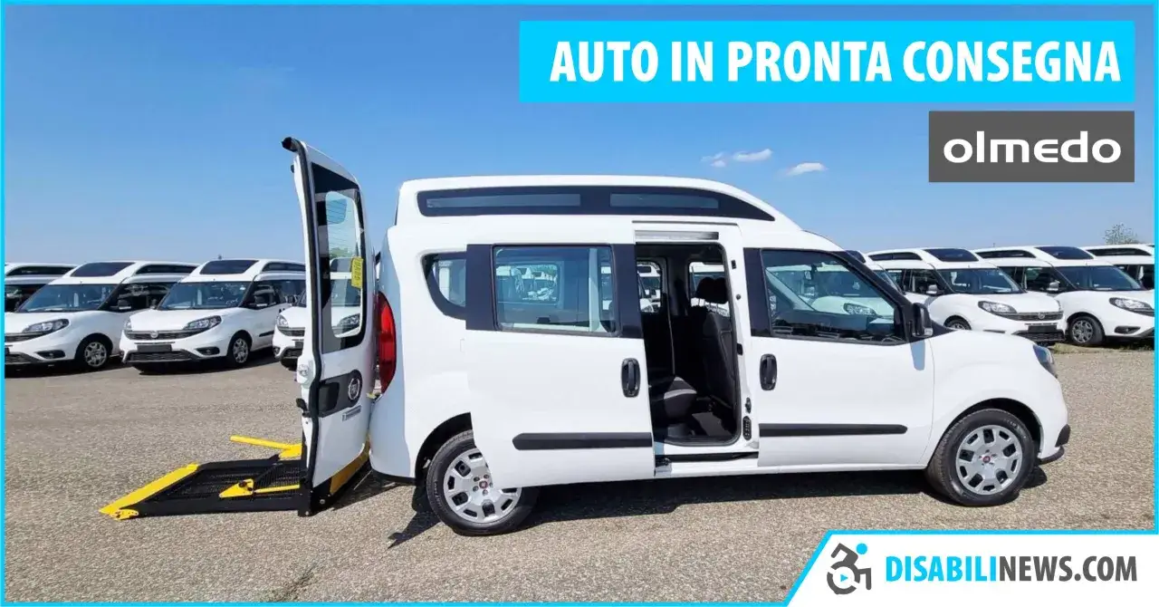 Auto bianca con bagagliaio per sedia a rotelle, pronta per la consegna. La rampa è estesa per facilitare l'accesso.