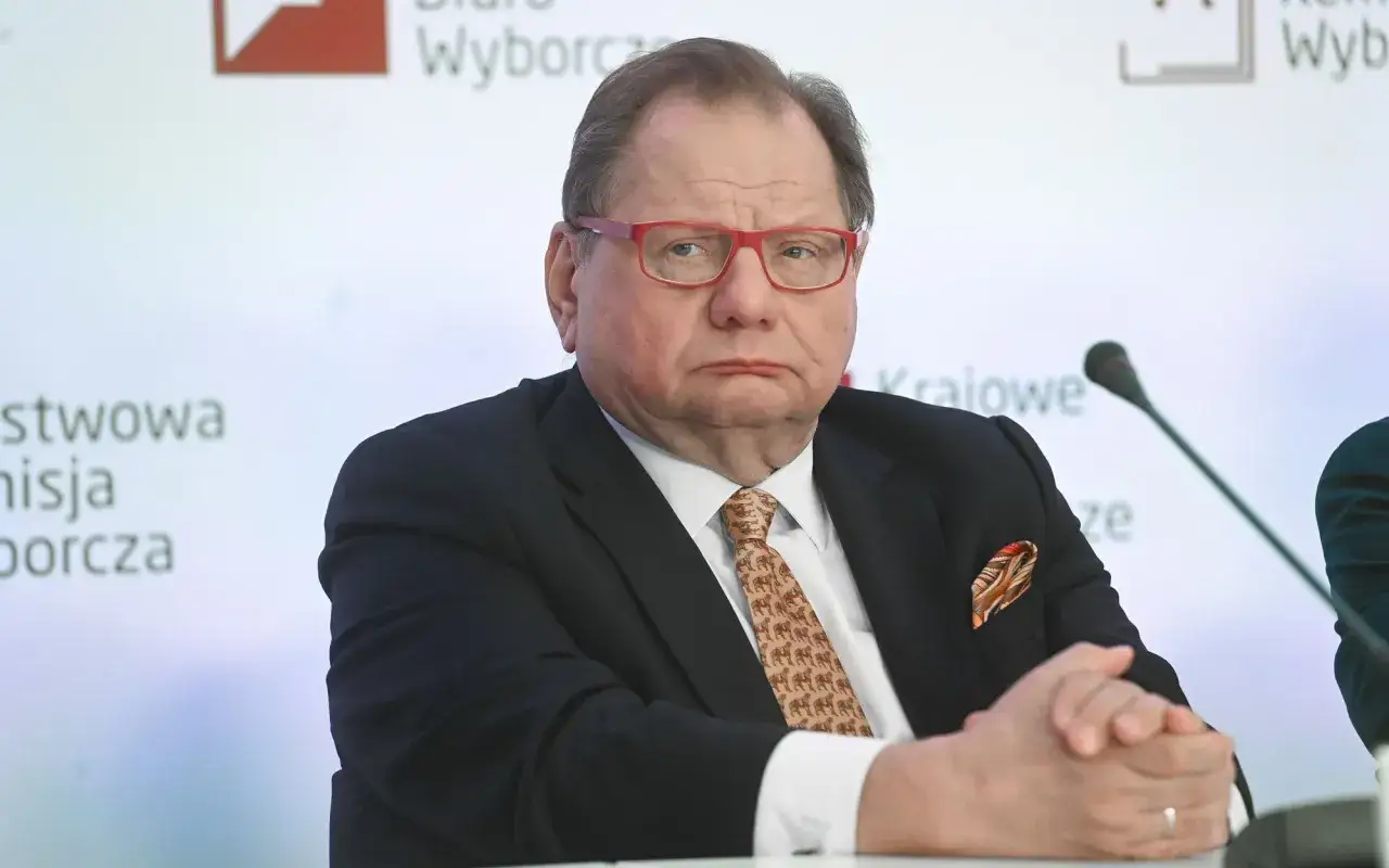 Mężczyzna w okularach i krawacie z wielbłądami, siedzi przy stole. Dyskutuje o biernym prawie wyborczym.