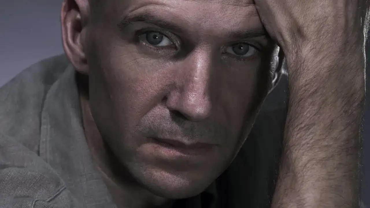 En närbild på Ralph Fiennes med handen mot pannan, hans blick är intensiv och tankfull.