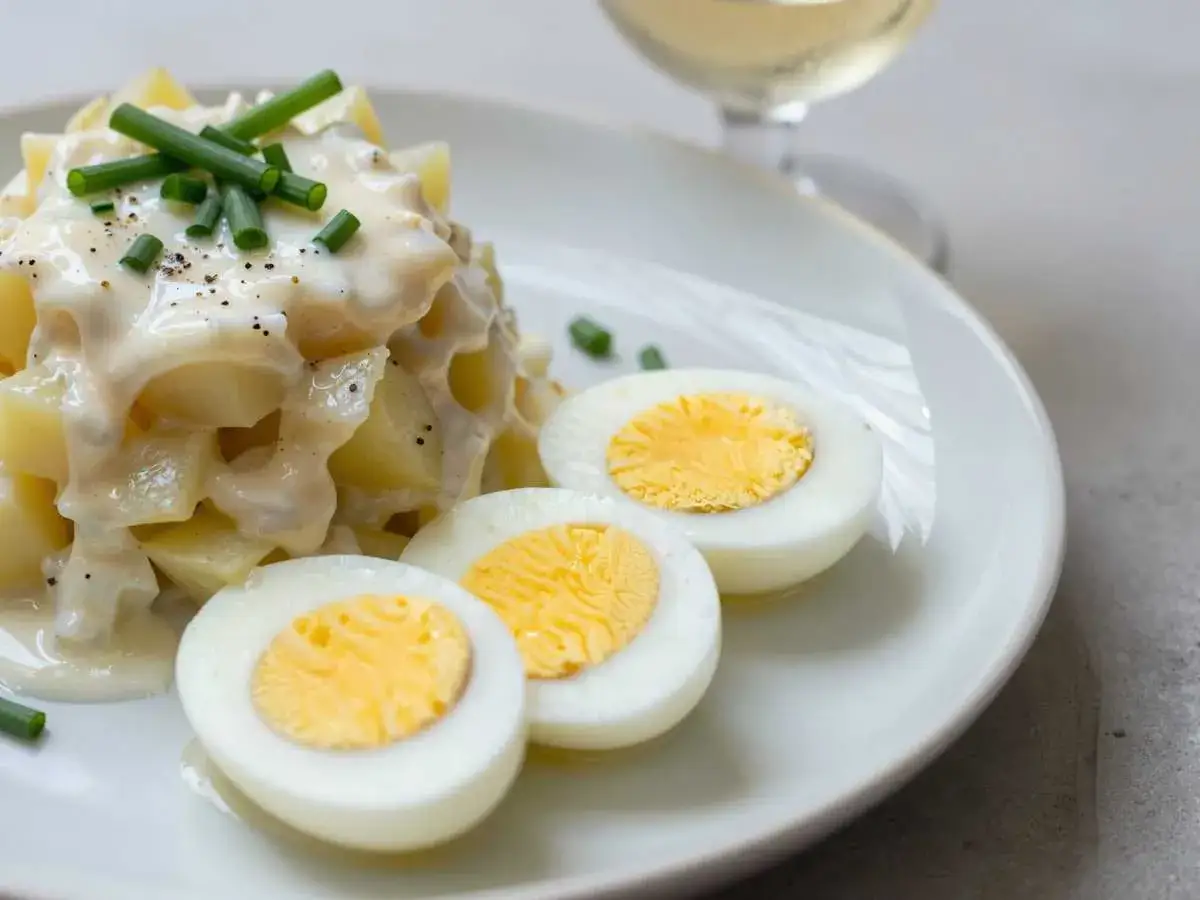 Cremiger Kartoffelsalat mit Gurken, garniert mit Schnittlauch und serviert mit halbierten gekochten Eiern.