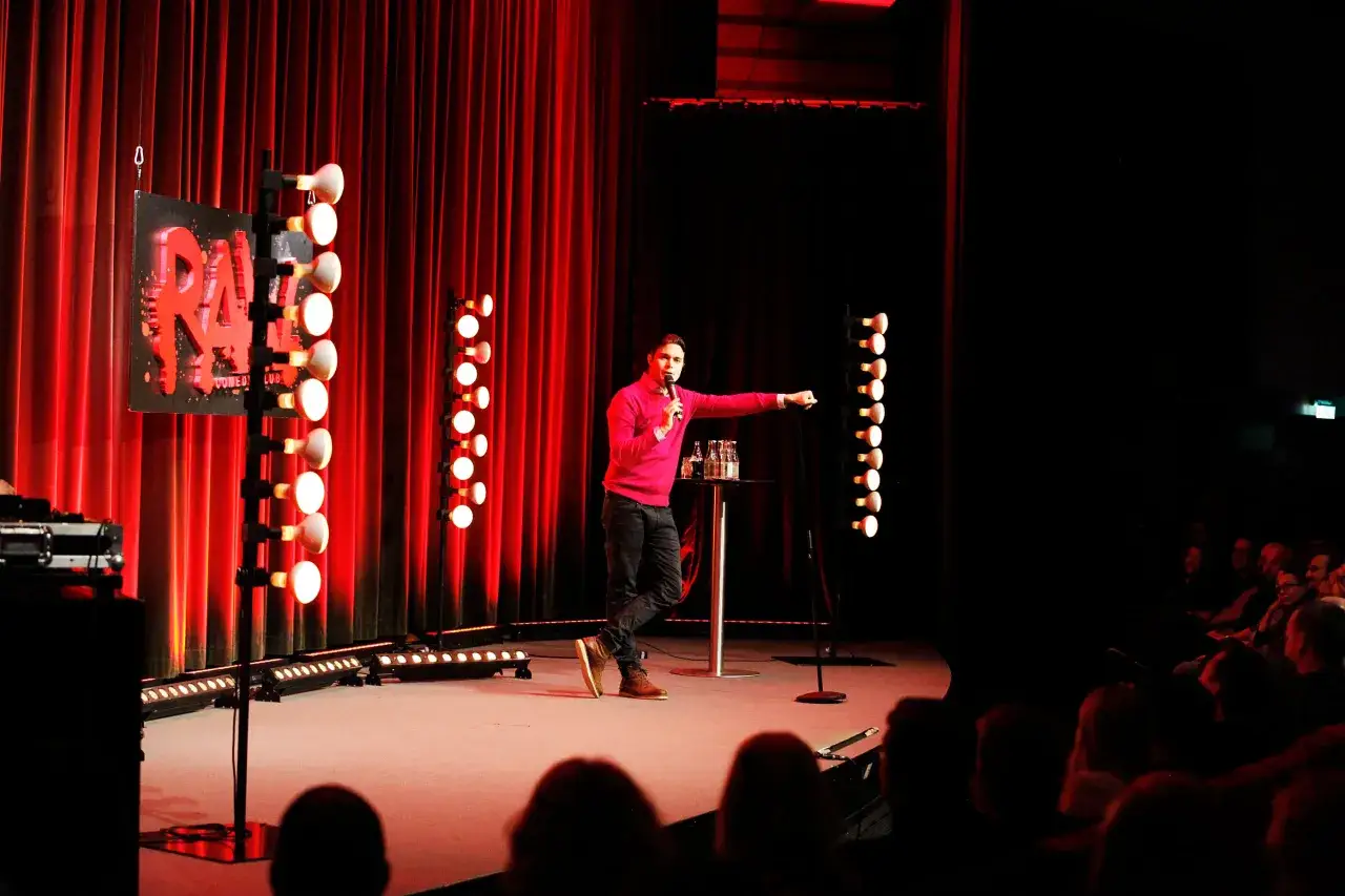 En stand up komiker i Sverige st&aring;r p&aring; scenen med en mikrofon. R&ouml;da draperier och lampor skapar en dramatisk st&auml;mning. Publiken sitter i m&ouml;rkret.