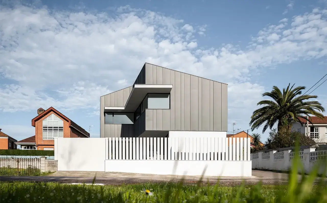 Moderna casa con **fachada zinc** gris, líneas angulosas y grandes ventanales, contrastando con una valla blanca y vegetación.