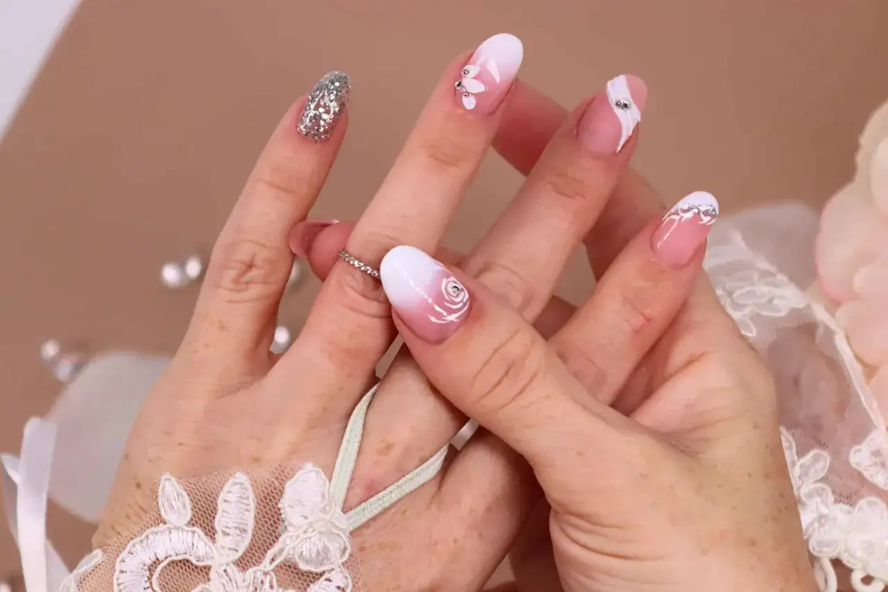 Manucure d&eacute;licate pour un mariage boh&egrave;me : ongles d&eacute;grad&eacute; blanc et rose, orn&eacute;s de roses, de strass et de paillettes argent&eacute;es.