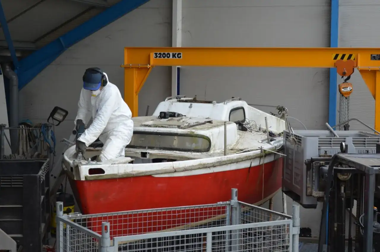 Un ouvrier en combinaison blanche travaille sur un bateau rouge et blanc, préparant son recyclage.