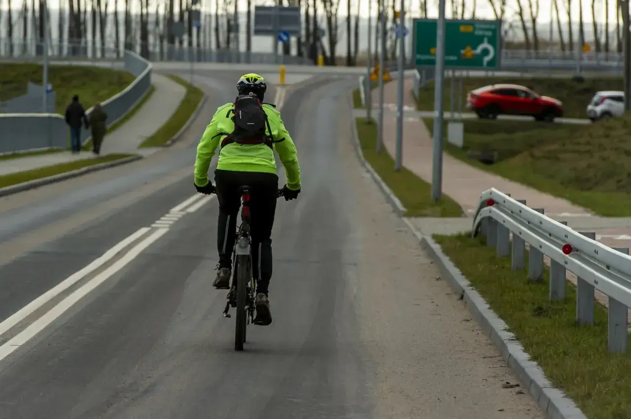 Kolarz w jaskrawej kurtce jedzie drogą. Czy rower to pojazd mechaniczny? Obok ścieżka dla pieszych i samochody.