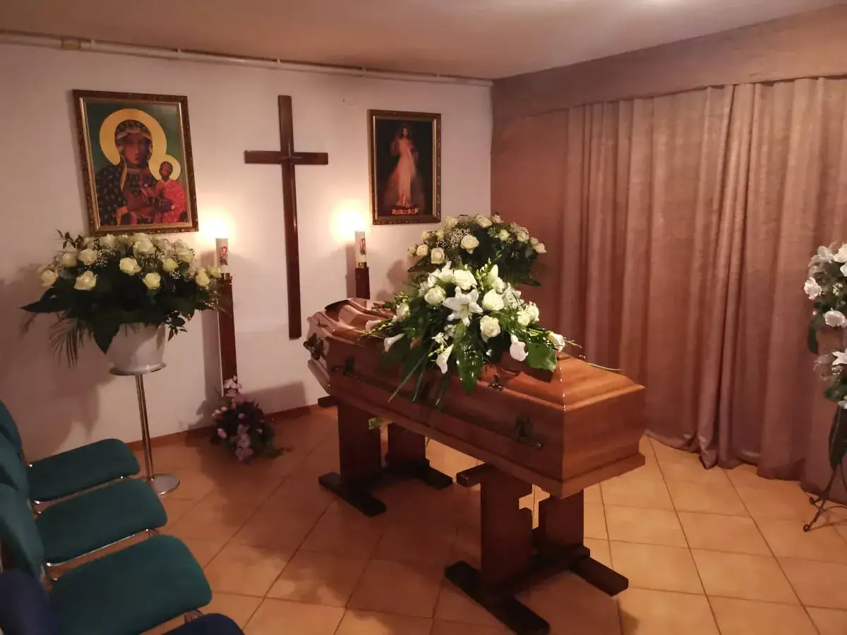 Przygotowania do chowania zmarłego po niedzieli. Drewniana trumna ozdobiona białymi kwiatami, obok obrazy religijne i krzyż.