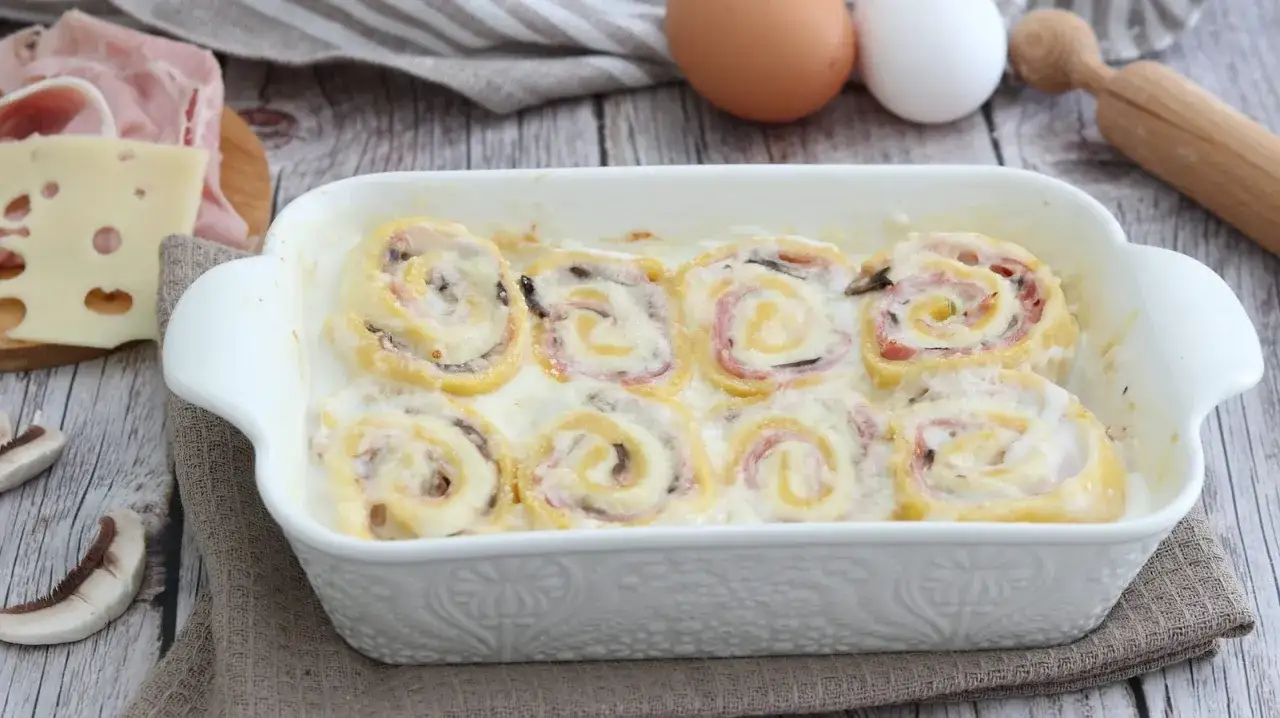 Nidi di rondine in una teglia bianca, ripieni di prosciutto e formaggio, con ingredienti freschi come uova e funghi sullo sfondo.
