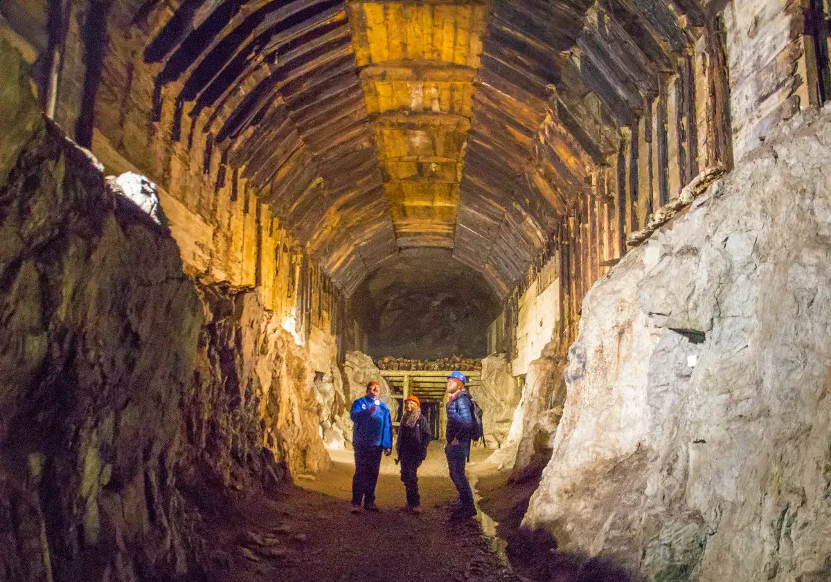 Trzech turystów zwiedza mroczny tunel, który jest częścią obiektu Riese. Drewniane wsporniki i skalne ściany tworzą surowy klimat.
