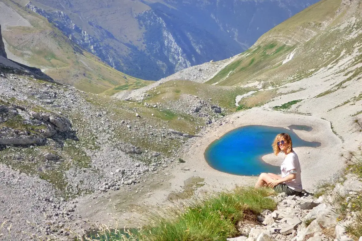 Donna seduta ammira il **lago di Pilato**, un gioiello blu incastonato tra montagne rocciose e verdi pendii.