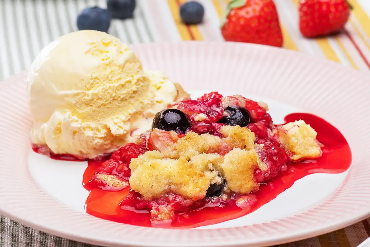Ein köstlicher Beeren Crumble mit einer Kugel Vanilleeis auf einem rosa gestreiften Teller.