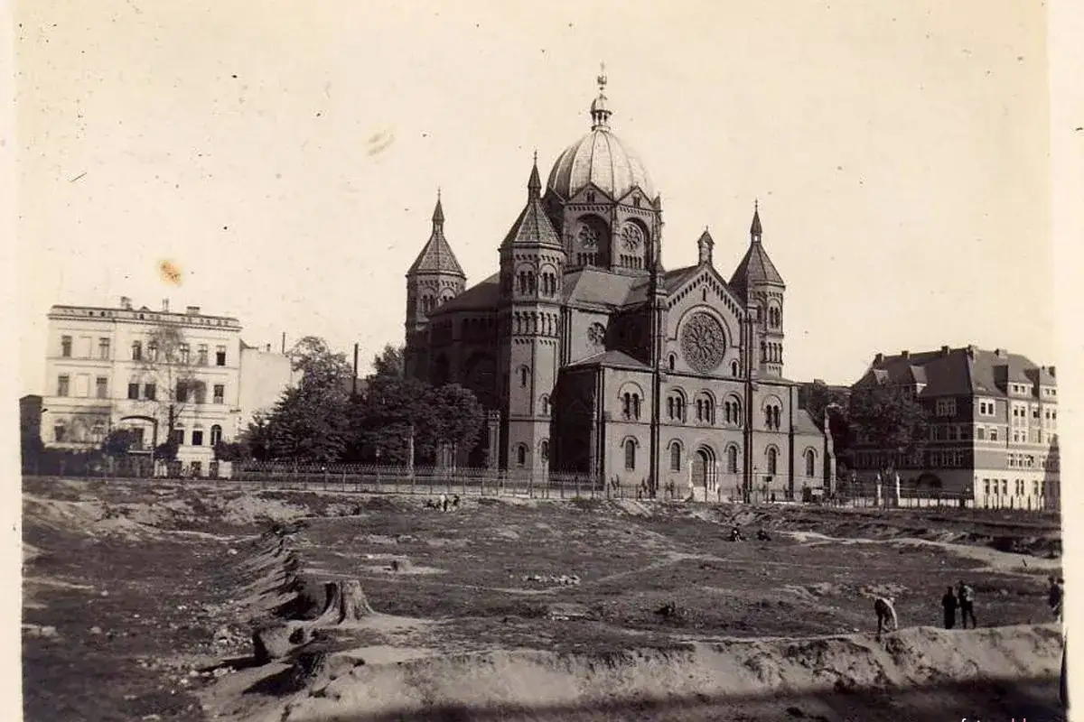 Imponująca synagoga z kopułą i wieżyczkami. W oddali budynki mieszkalne. Wok&oacute;ł plac budowy, gdzie dalej jest noc, a ludzie pracują.