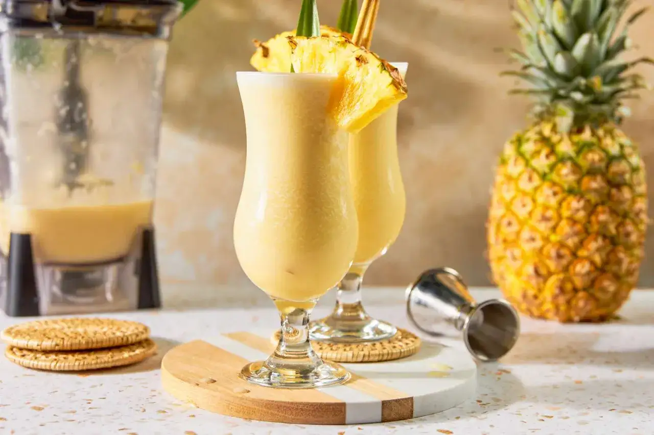 Deux verres de pina colada sans alcool, décorés de tranches d'ananas, prêts à être dégustés. Un ananas entier et un mixeur sont visibles en arrière-plan.