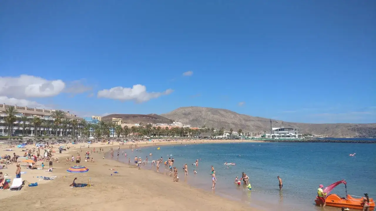 Słoneczny dzień na plaży Playa de las Américas. Ludzie kąpią się, opalają i bawią w wodzie. W tle hotele i góry.