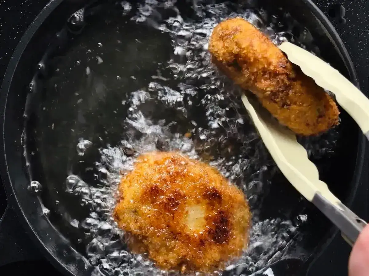 Golden potato croquettes are being fried in hot oil. One is held by tongs, ready to be flipped.