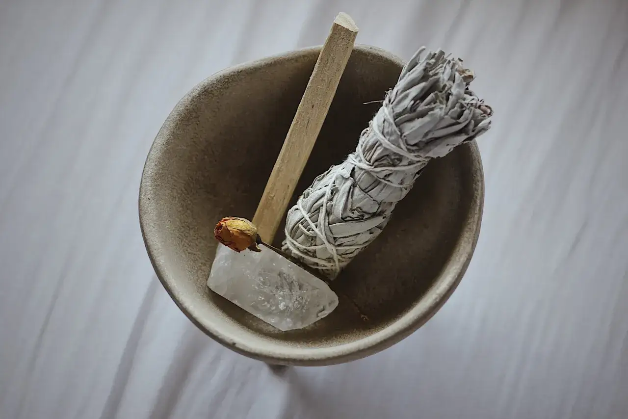 Un manojo de salvia blanca, un palo de palo santo, un cuarzo y una rosa seca en un cuenco. El incienso limpia casas para que sirve: purificar y armonizar espacios.