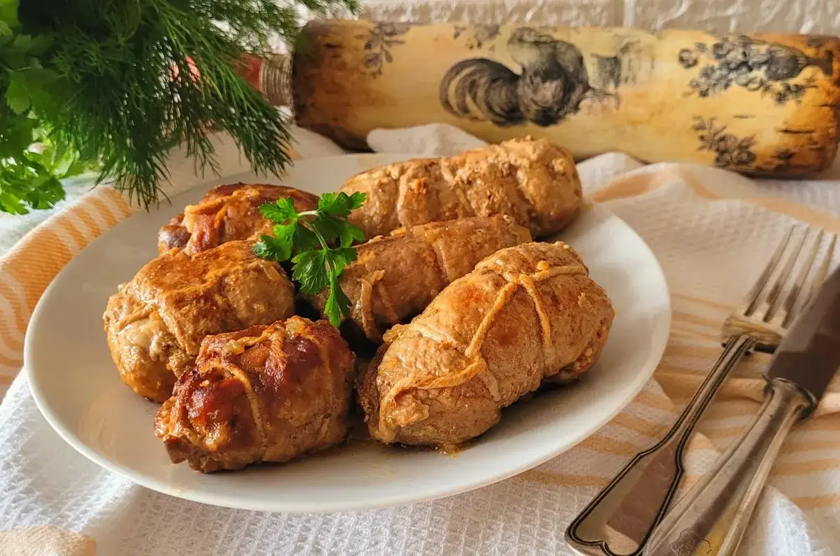 Zrazy z karkówki na białym talerzu, udekorowane natką pietruszki. Obok sztućce i dekoracyjny wałek do ciasta.