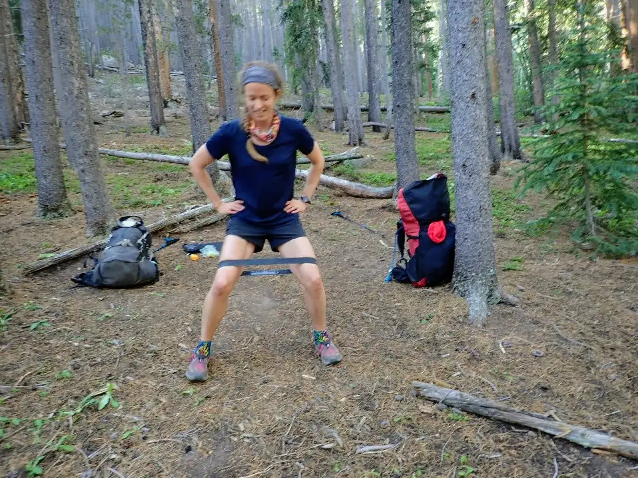 Femme en plein exercice &eacute;lastique dans une for&ecirc;t, avec des sacs &agrave; dos &agrave; proximit&eacute;.