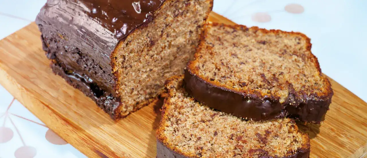 Saftiger Bananenkuchen mit Schokoladenglasur, der aussieht wie der beste Bananenkuchen der Welt. Zwei Scheiben liegen auf einem Holzbrett.