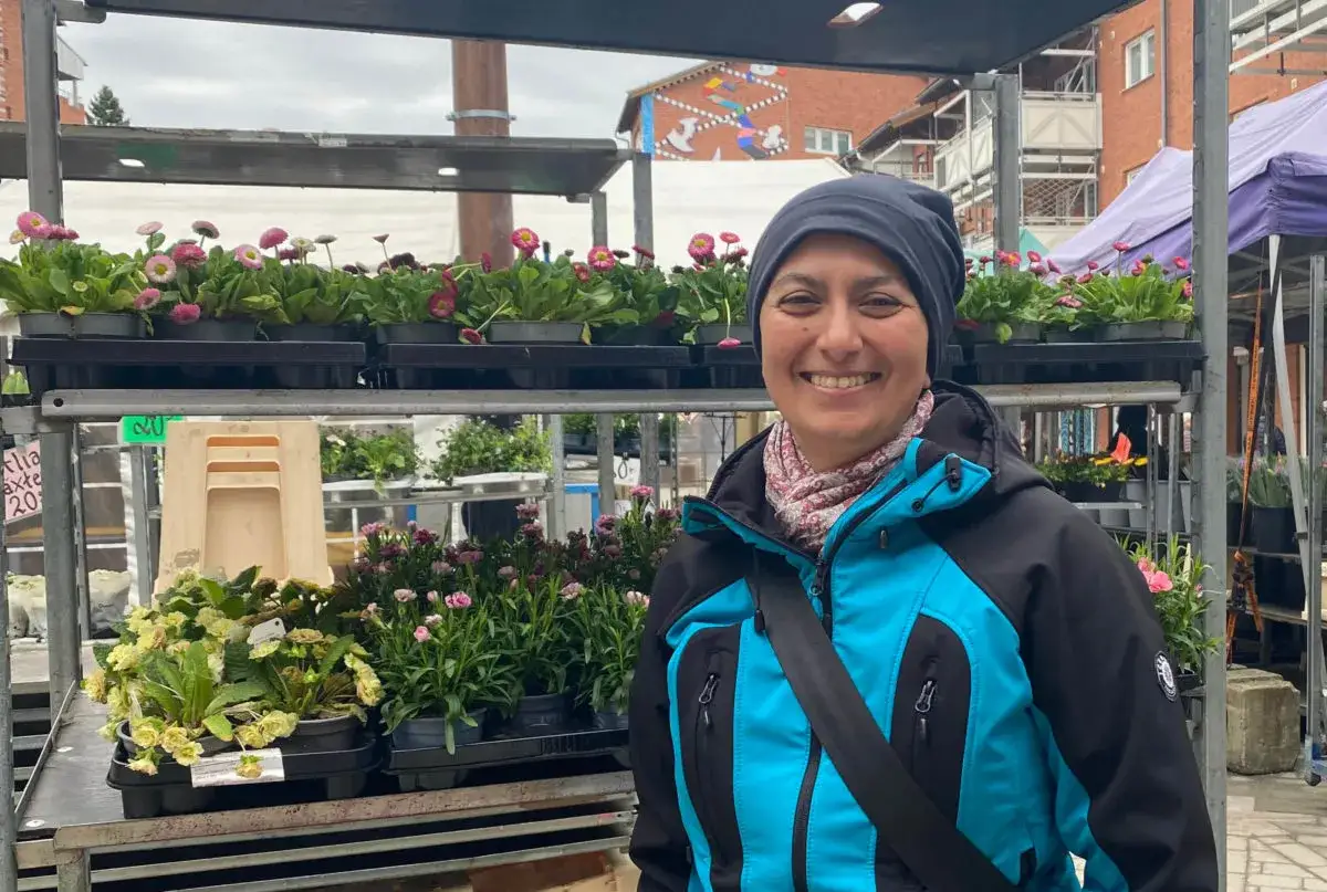 Zayera Khan ler vid ett blomst&aring;nd med rosa och vita tusensk&ouml;nor. Hon b&auml;r en bl&aring; jacka och en m&ouml;rk m&ouml;ssa.
