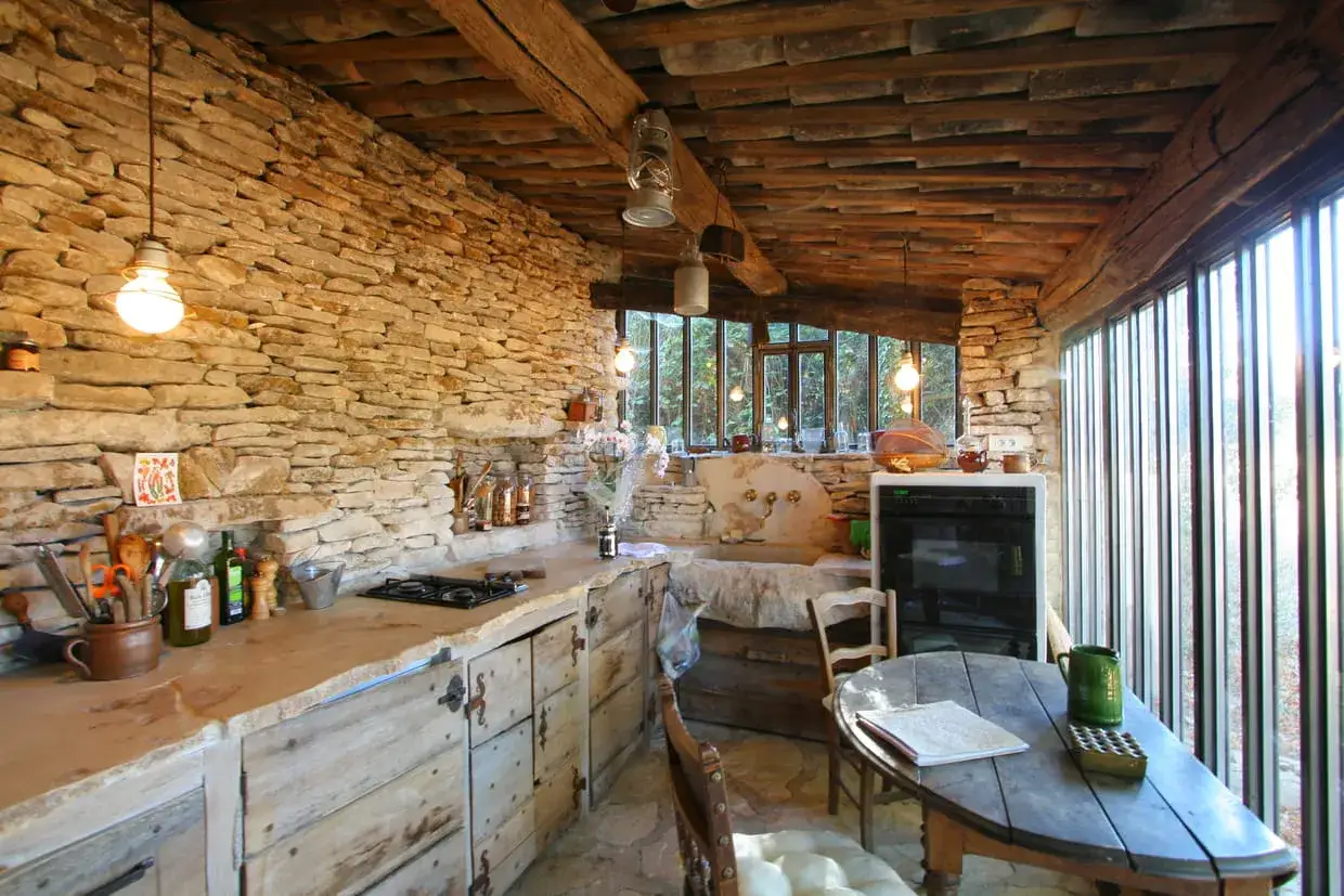 Cuisine rustique r&eacute;nov&eacute;e avec murs en pierre, plan de travail en pierre brute, meubles en bois vieilli et poutres apparentes.