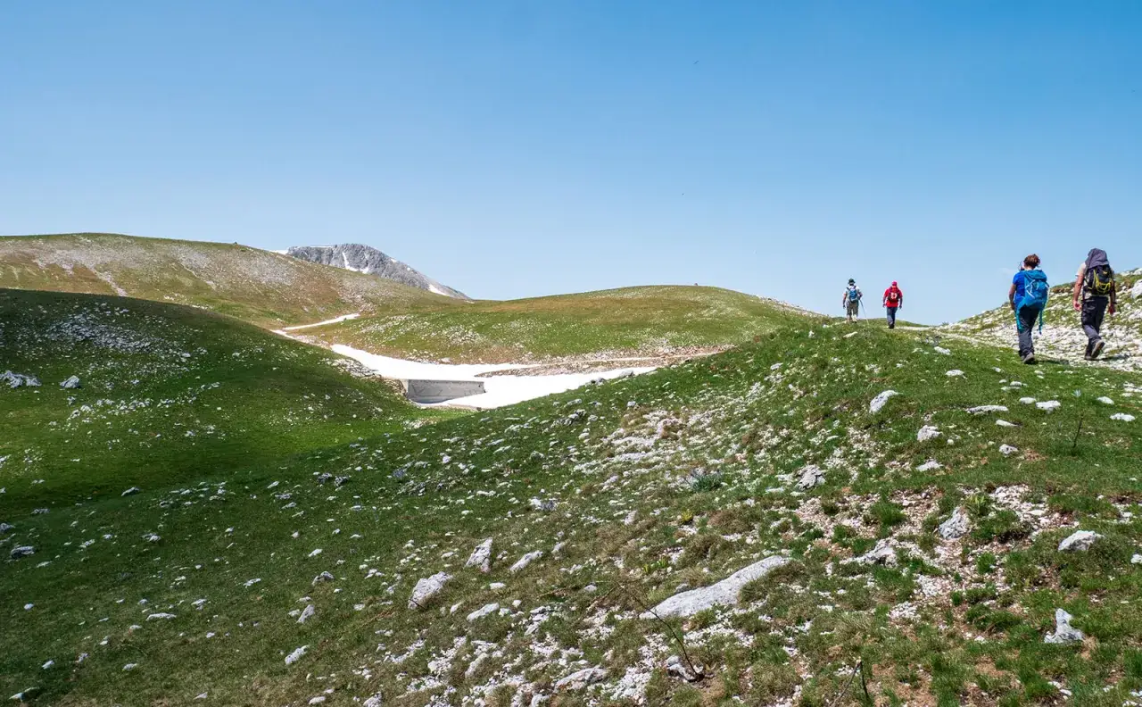 Escursionisti su un sentiero erboso con rocce, con neve residua e montagne sullo sfondo. L'aremogna Roccaraso altitudine offre panorami mozzafiato.