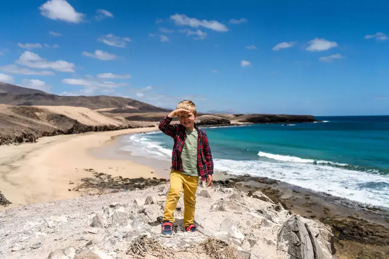 Niño sonriente en Lanzarote con niños, explorando una playa de arena dorada y aguas turquesas.