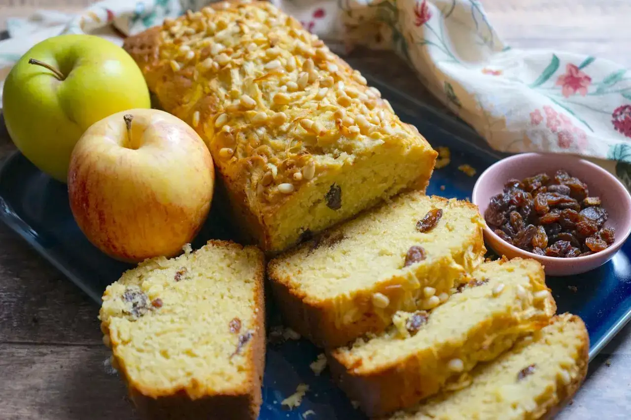 Plumcake alle mele dorato, cosparso di pinoli, con mele fresche e uvetta a lato.