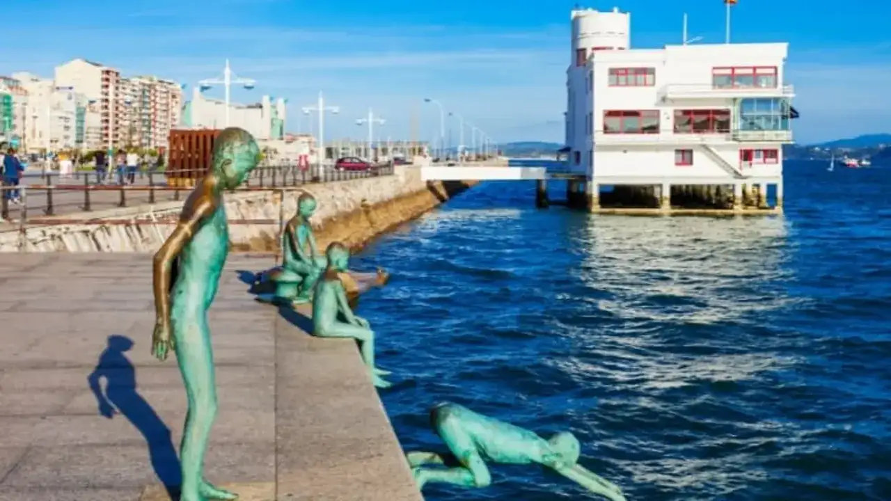 Las estatuas de los raqueros, niños jugando en el muelle, observan el mar azul junto a un edificio blanco sobre el agua.
