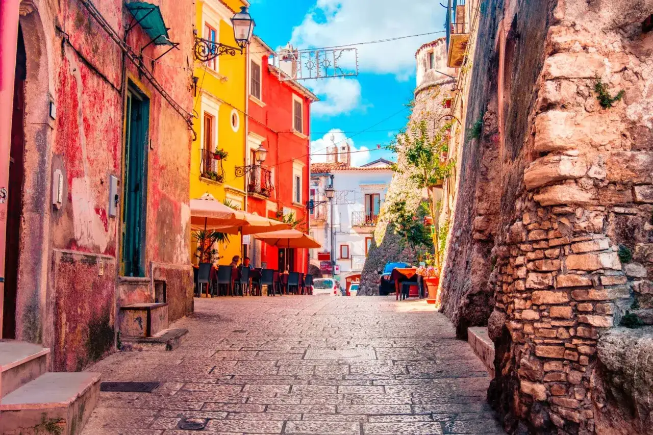 Un vicolo colorato a Vico del Gargano, con case vivaci, tavolini all'aperto e un'antica muraglia di pietra.