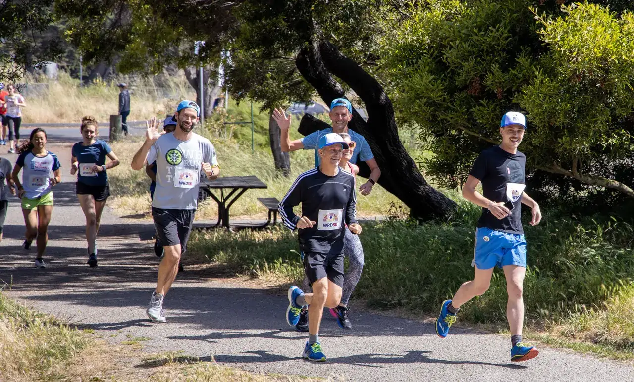 L&auml;ufer beim Rennen, einige mit Startnummern. Ein Mann winkt fr&ouml;hlich. Sie scheinen die 5 km in 20 min zu schaffen.