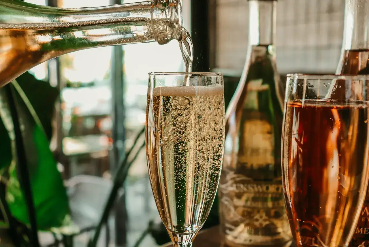 Nalewanie musującego drinka z prosecco do kieliszka, obok butelek i drugiego kieliszka z r&oacute;żowym napojem.