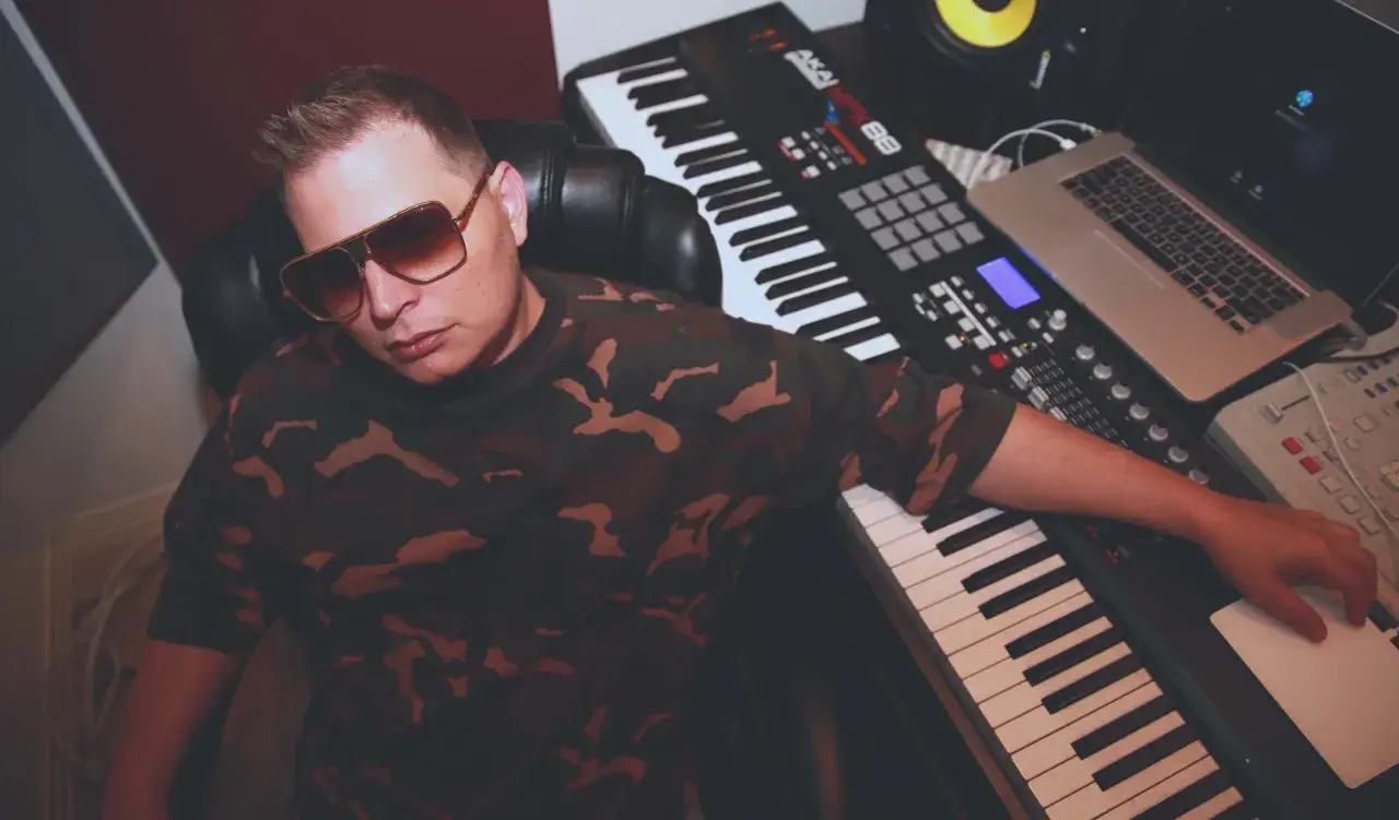 Scott Storch, wearing sunglasses and a camouflage shirt, sits in a studio surrounded by music equipment, ready to create more hit songs.