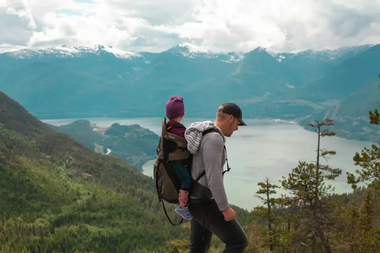 Ojciec z dzieckiem w nosidle podczas speed hiking po górach. W tle jezioro i ośnieżone szczyty.