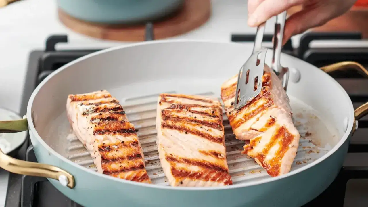 Drei saftige Lachsfilets braten in einer Grillpfanne mit schönen Grillstreifen. Eine Zange wendet gerade ein Stück.