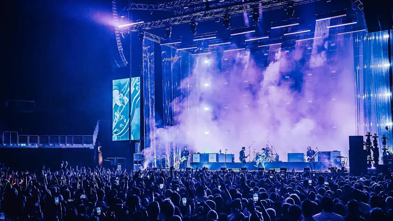 A band plays on a smoky stage under blue lights. The crowd cheers, holding up phones, as the screen behind them displays a guitar, hinting at the Nothing But Thieves setlist.