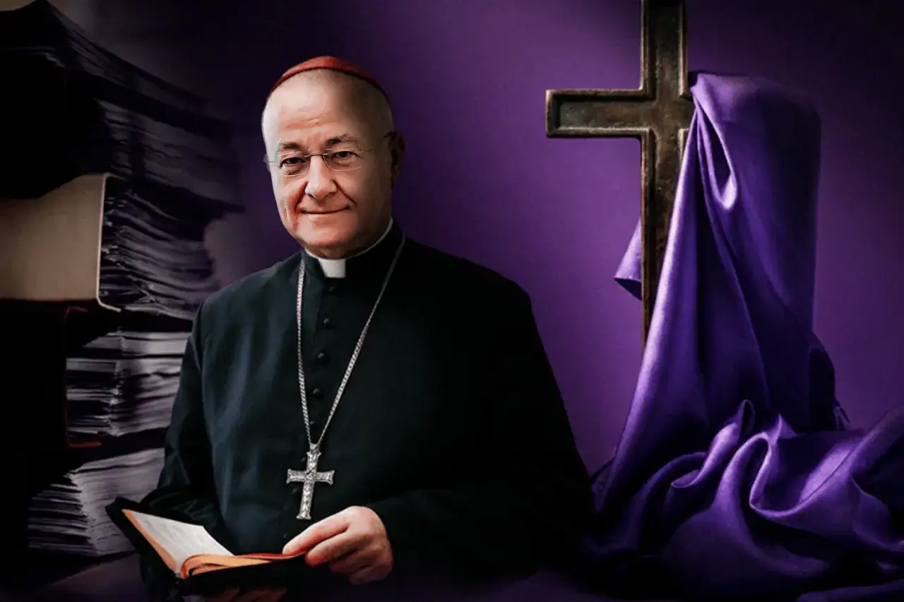 Uśmiechnięty biskup w czarnej sutannie z krzyżem na łańcuchu, trzyma otwartą księgę. W tle stos dokumentów i krzyż okryty fioletową tkaniną.