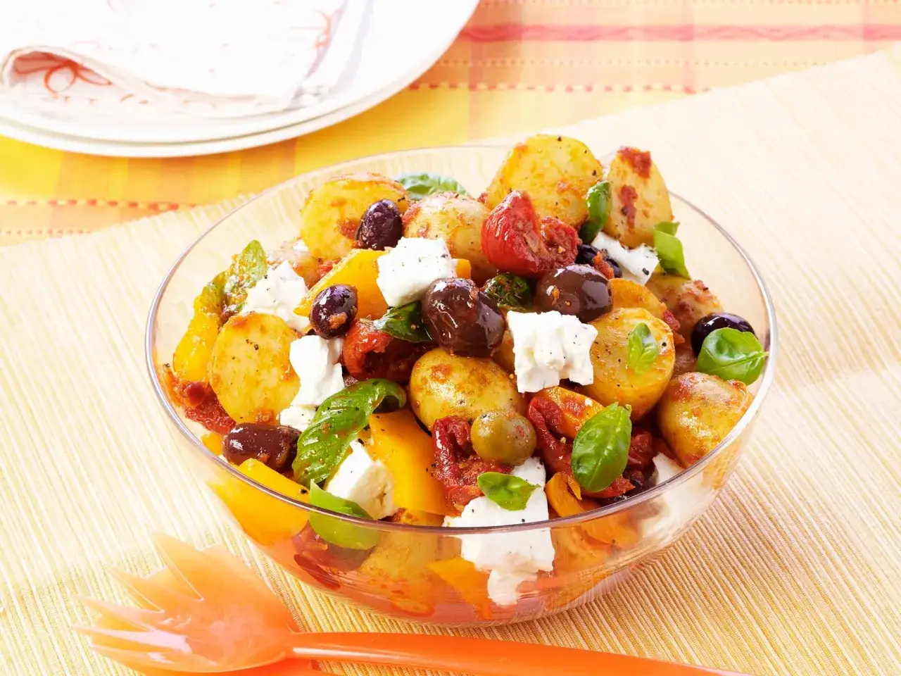 Ein bunter mediterraner Kartoffelsalat mit Oliven, Tomaten, Paprika und Feta in einer Glasschüssel.