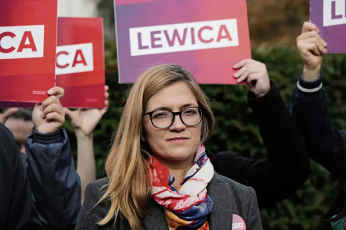 Kobieta w okularach i kolorowym szaliku, na tle osób trzymających czerwone i fioletowe tabliczki z napisem "LEWICA".