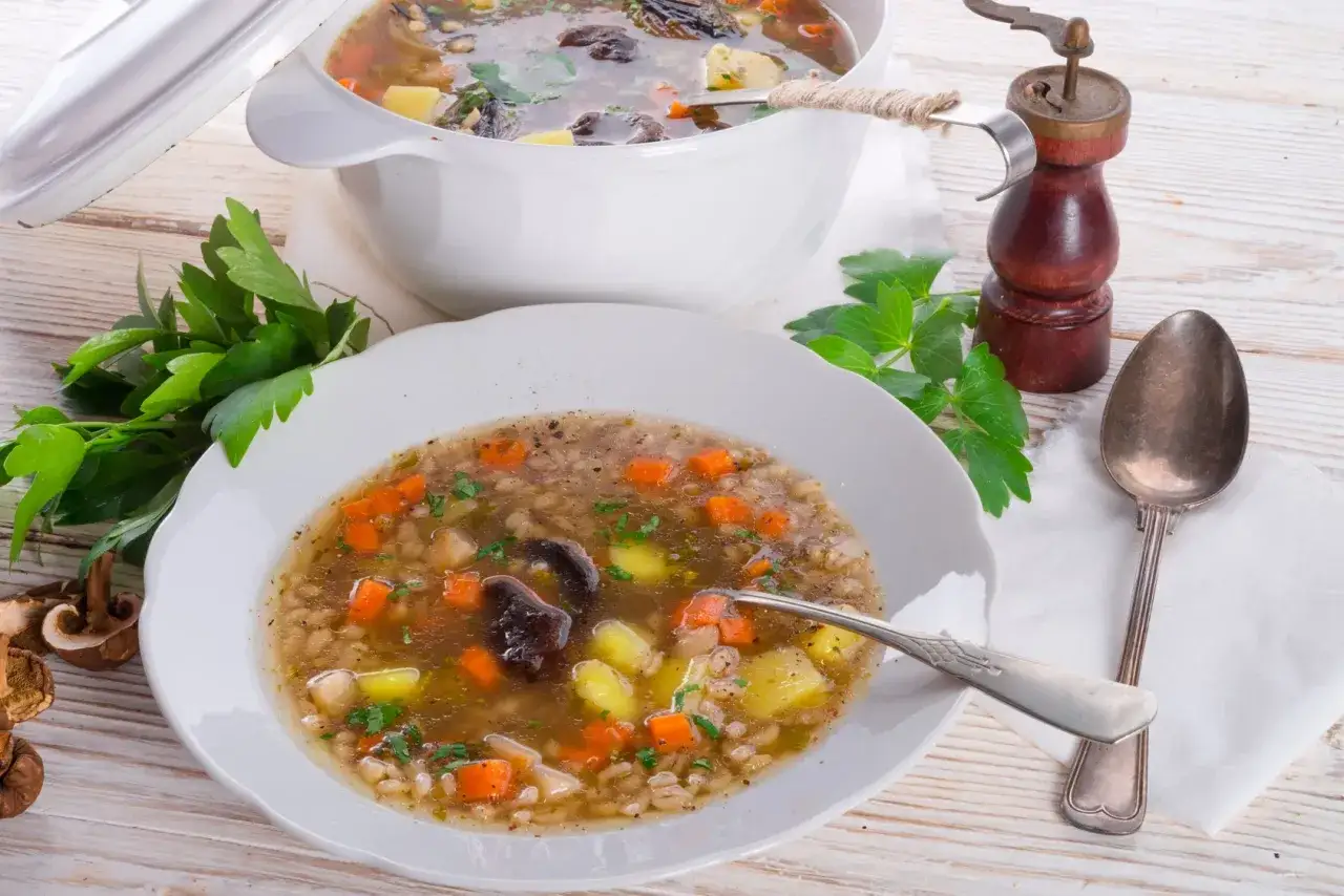 Aromatyczna zupa krupnik Magdy Gessler z grzybami, ziemniakami i marchewką. Podana w białej misce, obok garnek z zupą.
