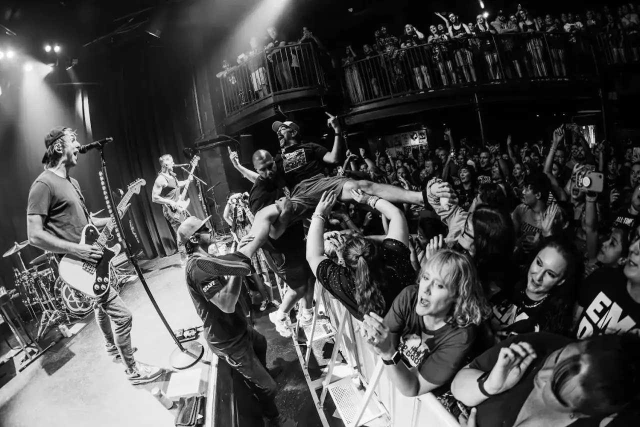 Energetyczny koncert, tłum szaleje, a na scenie zespół grający w stylu emo. Widać gitarzystę, basistę i wokalistę, a przed sceną fani unoszą jednego z członków zespołu.