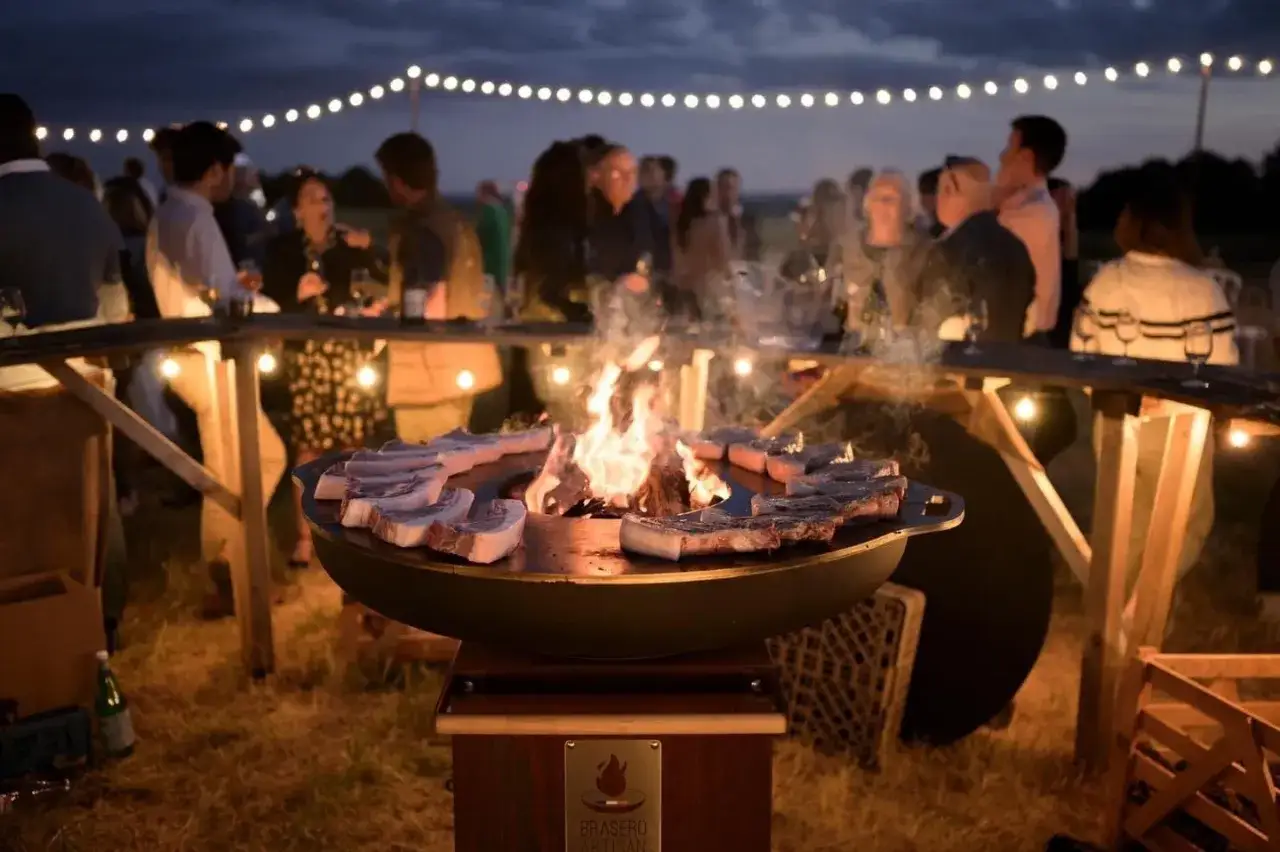 Un barbecue pour mariage en plein air, avec des côtelettes sur le feu et des invités profitant de la soirée sous des guirlandes lumineuses.