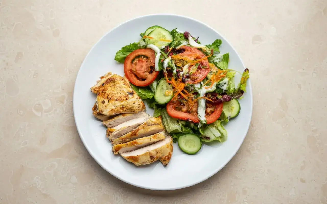 Deliciosa ensalada de pollo con lechuga, tomate, pepino y zanahoria rallada, acompañada de pechuga de pollo a la parrilla en rodajas.