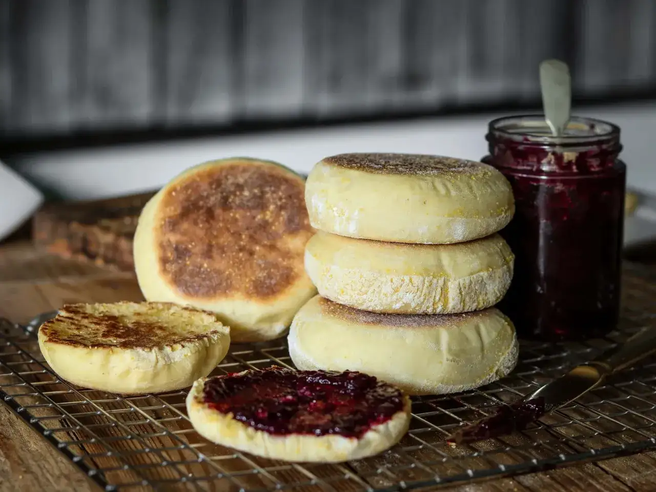 Ein Stapel goldbrauner englischer Muffins, einer davon mit Marmelade bestrichen, neben einem Glas Beerenmarmelade.
