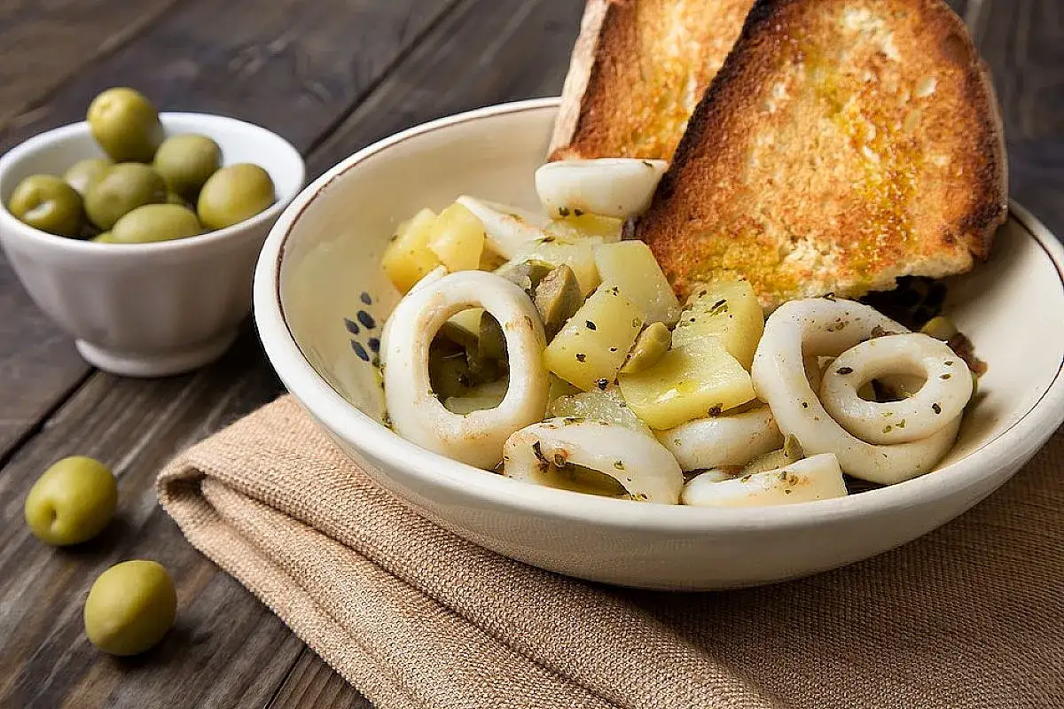 Anelli di totano in padella con patate e olive, accompagnati da pane tostato e un contorno di olive verdi.