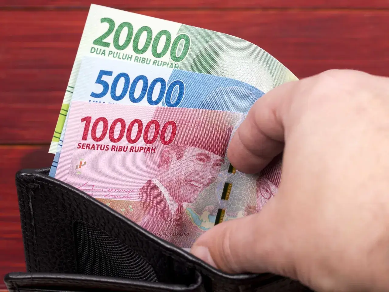 Ręka wsuwa do portfela banknoty: 20 000, 50 000 i 100 000 rupii. To waluta na Bali, idealna na wakacyjne wydatki.
