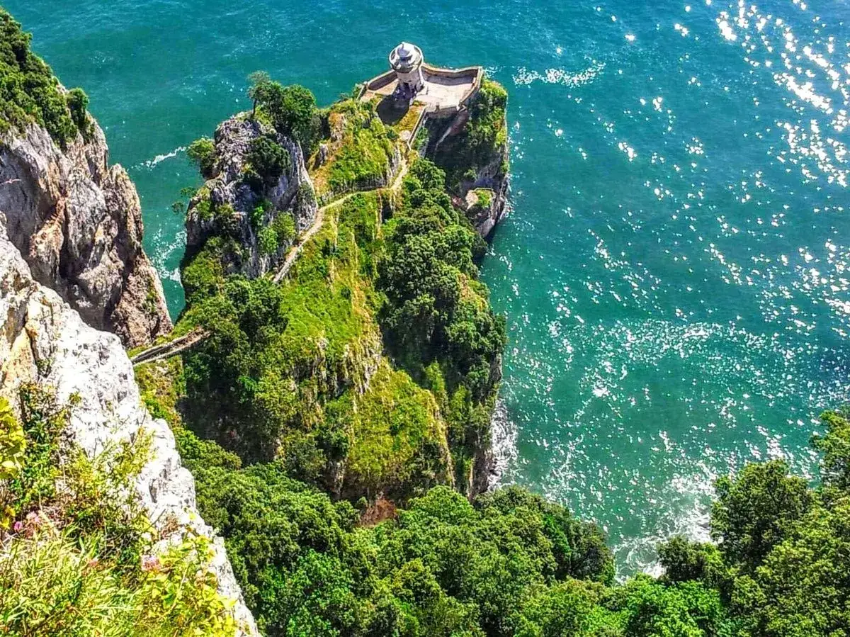 Un faro blanco se alza en un acantilado verde, con un camino sinuoso que lleva hasta él. El mar turquesa brilla bajo el sol. Este cabo de ajo es un lugar de ensueño.