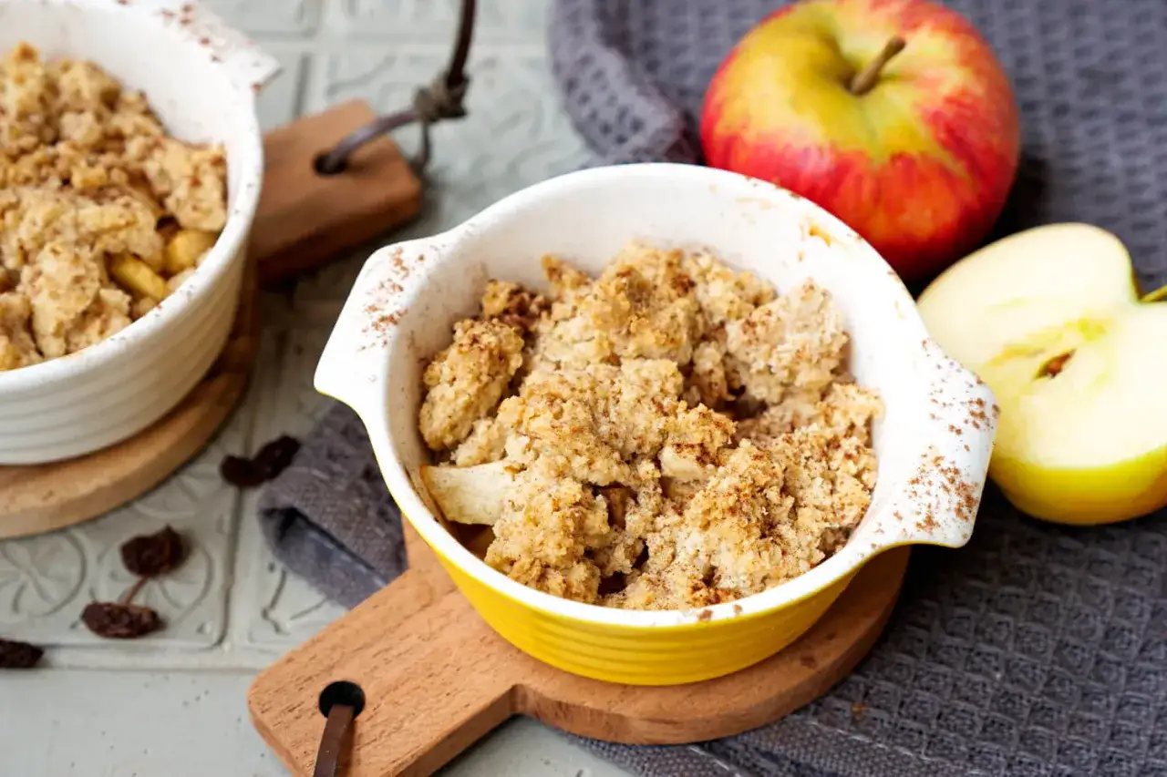 Zwei Schalen mit warmem Apfel Crumble mit Haferflocken, daneben ein ganzer und ein halber Apfel.