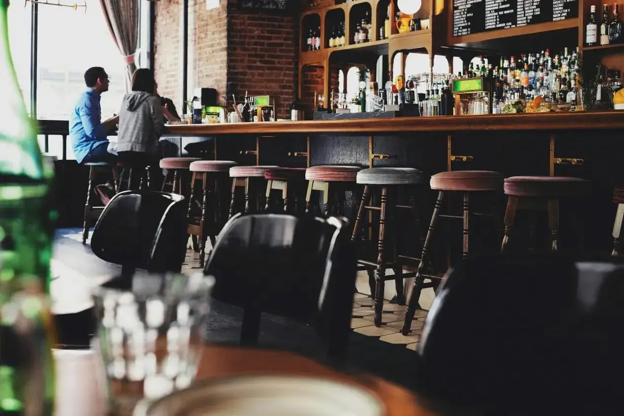 Bar con taburetes y estantes llenos de botellas. Dos personas sentadas en la barra, disfrutando de bebidas. Un lugar ideal para una licencia de bar sin cocina.