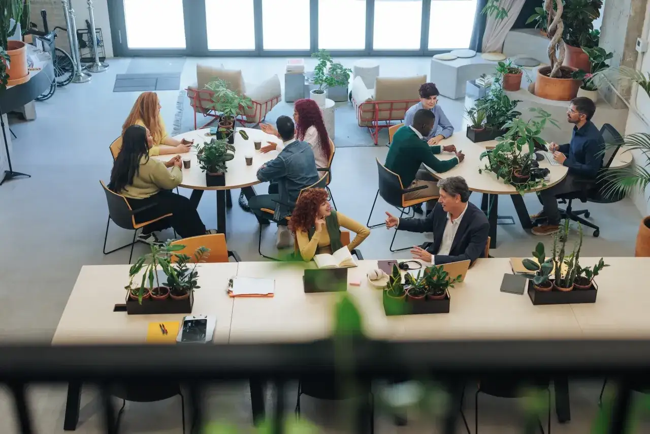 Ludzie pracują i rozmawiają w jasnej przestrzeni, która pokazuje, coworking co to: elastyczne biuro pełne zieleni i współpracy.