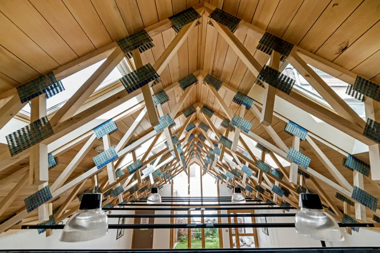 Vue en contre-plong&eacute;e d'une charpente sans ferme, avec des poutres en bois et des connecteurs bleus. Des luminaires industriels pendent sous la structure.